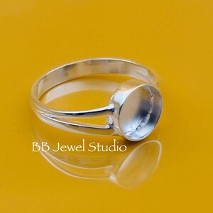 May include: A silver ring with an oval setting, designed to hold a gemstone. The ring has a split shank design and is displayed on a yellow surface. The text "BB Jewel Studio" is visible.