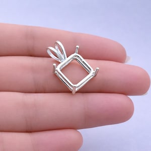 May include: A silver pendant setting with a square shape and four prongs. The pendant has a loop for a chain and is designed to hold a gemstone. The setting is shown against a light purple background.