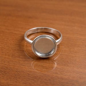May include: A silver ring with a round bezel setting. The ring has a simple band and a circular setting designed to hold a stone or other embellishment. The ring is displayed on a wooden surface.