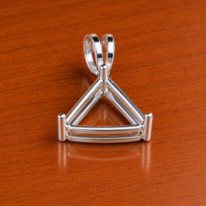 Op de afbeelding: Een gepolijste zilverkleurige metalen hangerzetting met een driehoekig frame ontworpen om een edelsteen vast te houden. De zetting bevat drie verticale pootjes op de hoeken en een stevig, afgerond oogje aan de bovenkant voor een ketting.