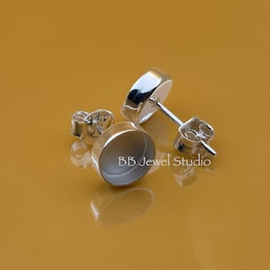 May include: Close-up of three silver stud earrings with posts and backs. The earrings have a round, bezel setting. The text "BB Jewel Studio" is visible on the surface. The background is a solid, warm, golden-brown color.