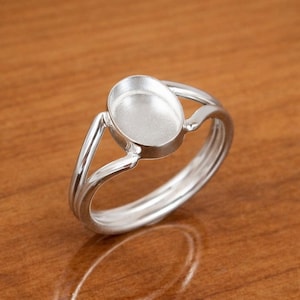 Könnte beinhalten: Ein silberfarbener Ring mit ovaler Fassung. Der Ring hat ein geteiltes Schaftdesign mit zwei Bändern, die sich in der Nähe der Fassung treffen. Die Fassung ist für einen Stein oder ein anderes dekoratives Element vorgesehen. Der Ring wird auf einer Holzoberfläche gezeigt.
