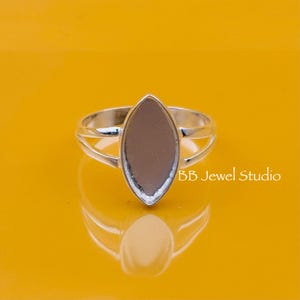 May include: A silver ring with a large, oval-shaped setting. The setting is a light brown color and is surrounded by a silver band. The ring is set against a yellow background. The text "BB Jewel Studio" is visible.