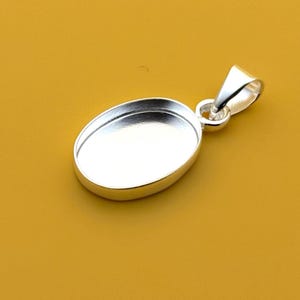 Op de afbeelding: Een zilverkleurige ovale hanger met een lus voor een ketting. De hanger heeft een verzonken gebied voor het plaatsen van een steen of ander decoratief element. De achtergrond is effen mosterdgeel.