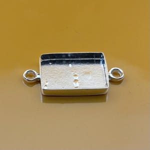 May include: A rectangular silver-coloured jewellery finding with a recessed area and two small loops on either side. The finding is designed to hold a stone or other decorative element. The background is a solid, warm yellow colour.