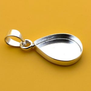 May include: A silver-colored pendant with a teardrop-shaped bezel. The pendant has a loop at the top for a chain and a recessed area to hold a stone or other decorative element. The pendant is displayed against a yellow background.