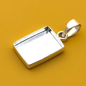 Puede incluir: Un colgante rectangular plateado con un lazo para una cadena. El colgante tiene un área empotrada, probablemente para engarzar una piedra u otro elemento decorativo. El colgante se muestra sobre un fondo amarillo.