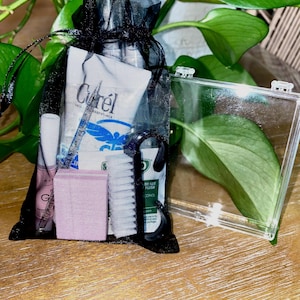 May include: A black mesh drawstring bag filled with travel-sized toiletries, including a tube of CeraVe lotion, a nail file, and a small pink buffer. A clear plastic box sits to the right. The items are on a wooden surface with green plants in the background.