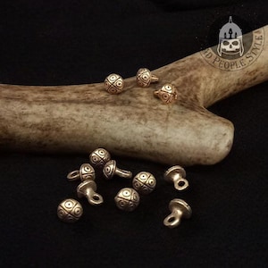 Può includere: Una collezione di piccoli bottoni metallici rotondi con un motivo a croce e punti sulla superficie. Ogni bottone ha un occhiello sul retro per l'attacco. I bottoni hanno una finitura metallica e sono disposti in un gruppo.