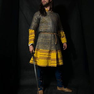 May include: A medieval-style costume featuring a chainmail shirt with yellow and brown accents, a matching skirt, and blue pants. The person is holding a sword and wearing brown leather shoes. The background is dark.