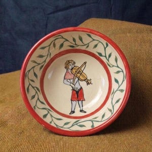 May include: A round ceramic bowl with a red rim and a central illustration of a person playing a violin. The bowl's interior features a red band and a green vine pattern around the edge. The person is dressed in red and white clothing.