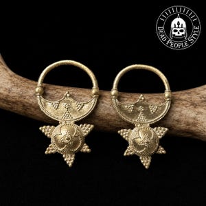 Op de afbeelding: Een paar goudkleurige metalen oorbellen met een cirkelvormige ring aan de bovenkant, verbonden met een halvemaanvormig stuk met textuurdetails, eindigend in een stervormige hanger met een centrale koepel en kralenranden. Logo is zichtbaar.