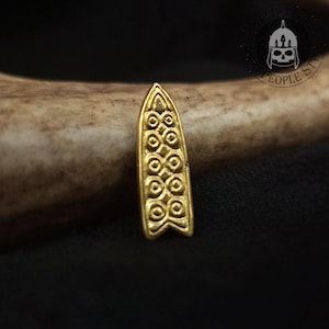 Puede incluir: Un colgante de metal dorado con forma de escudo puntiagudo o tabla de surf, que presenta un patrón en relieve repetitivo de círculos y líneas interconectadas. El objeto tiene un acabado metálico liso y un diseño geométrico simétrico.