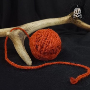May include: A ball of orange yarn with a loose strand extending to the left. The yarn is resting on a dark surface, with a piece of antler in the background. The yarn appears to be made of a natural fiber.