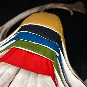 Puede incluir: Un artículo textil multicapa con una parte superior amarilla, capas negras, azules, verdes y rojas, y un interior acolchado blanco. Las capas están separadas por un ribete blanco. Una cornamenta curvada en el fondo.
