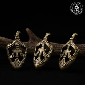 Puede incluir: Tres colgantes de metal en tono latón antiguo con forma de escudo y diseño calado que presenta un pilar central vertical y detalles curvos. Cada pieza tiene una pequeña anilla en la parte superior y un acabado texturizado y desgastado.
