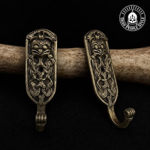 Op de afbeelding: Twee antiek messingkleurige metalen wandhaken met gedetailleerde reliëfsculpturen van een gestileerd wezen met grote ogen en verweven knooppatronen. Elke haak heeft een gebogen onderkant en een rechthoekig bovendeel met een montagegat.