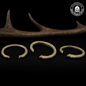 Könnte beinhalten: Drei offene Armreifen aus Messing mit einer strukturierten, schuppenartigen Oberfläche und stilisierten Tierkopf-Endstücken. Das Metall hat einen matten Goldton und jedes Stück weist ein geschwungenes Design mit feinen Details auf.