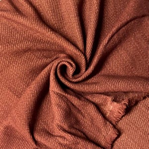 May include: Close-up of a folded, rust-coloured fabric with a textured weave. The fabric is bunched and swirled, creating deep shadows and highlights. The edges of the fabric are frayed.