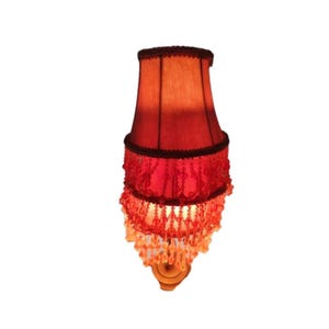 Puede incluir: Una pantalla de lámpara roja y naranja con flecos de cuentas. La pantalla tiene un ribete marrón oscuro y una base de madera. La sombra es de color rojo intenso en la parte inferior, desvaneciéndose a naranja en la parte superior. Los flecos son de cuentas transparentes y naranjas.