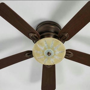 May include: Ceiling fan with five dark brown wooden blades. The fan's motor housing is a bronze color, and the light fixture has a yellow and blue floral design. The fan is mounted on a white ceiling.