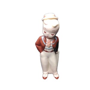 May include: A vintage ceramic figurine of a boy in a brown jacket, white pants, and a small hat. The figurine has a painted face with closed eyes and red accents on the shoes. The boy is holding a white object.