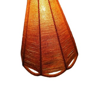 Puede incluir: Primer plano de una pantalla de lámpara naranja hecha a mano. La pantalla está hecha de material tejido, con forma cónica y una base abierta y decorativa. La textura del material tejido es visible, y la pantalla está iluminada desde dentro.