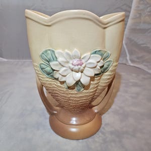 May include: A beige ceramic vase with a decorative flower design. The vase features a textured basket weave pattern and a sculpted white flower with a pink center and green leaves. The vase has a rounded base and two handles.