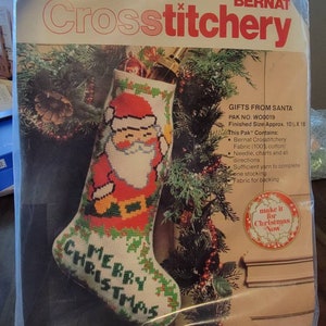 May include: A cross stitch kit featuring a red and white stocking with a Santa Claus design and the words "Merry Christmas". The kit includes yarn, fabric, a needle, and instructions. The packaging is labelled "Gifts from Santa" and "Bernat Crosstitchery".