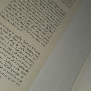 Może przedstawiać: Otwarta książka z tytułem "Talking to the Dead" i nazwiskiem autora. Okładka zawiera logo z literą "M". Strony są w kolorze ecru, a tekst jest czarny.