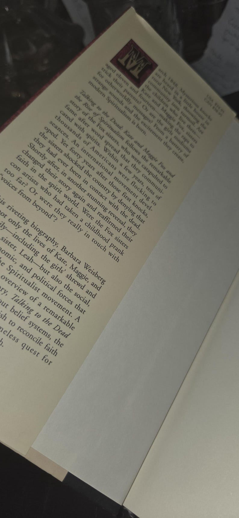 Może przedstawiać: Otwarta książka z tytułem "Talking to the Dead" i nazwiskiem autora. Okładka zawiera logo z literą "M". Strony są w kolorze ecru, a tekst jest czarny.