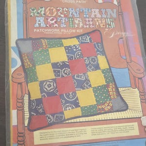 May include: A patchwork pillow kit in a clear plastic package. The kit includes a pillow with a colorful patchwork design of squares in red, yellow, blue, and green. The packaging features text that reads "Mountain Artisans Patchwork Pillow Kit."