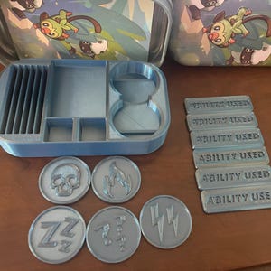 May include: A light blue 3D-printed organizer for trading card game accessories. Includes a card holder, dice holders, and coin holders. Also shown are game tokens with various symbols and "ABILITY USED" markers.