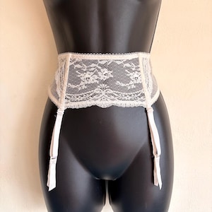 May include: A white lace garter belt featuring a wide, intricate floral pattern across the waistband. The garment includes four adjustable white elastic straps with metal clips hanging from the bottom edge to secure hosiery in place.