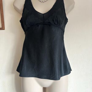 May include: Black camisole top with a V-neck and a slightly flared hem. The top has a delicate, textured trim along the neckline and straps. A silver necklace with small, round embellishments is visible. The top is displayed on a mannequin.