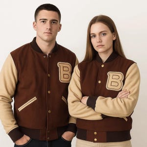 May include: Two brown varsity jackets with beige sleeves and a large letter "B" patch. The jackets have a button-up front, a ribbed collar, and cuffs. The jackets are worn by two people.