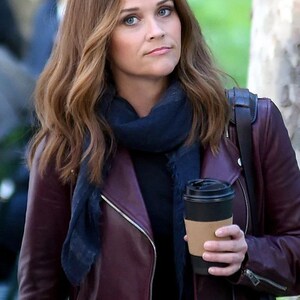 May include: A burgundy leather jacket with a black top and a navy blue scarf. The person is holding a black coffee cup with a brown sleeve. The jacket has a zipper and a black belt.