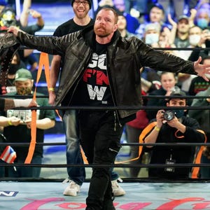 Op de afbeelding: Een man in een zwarte leren jas en zwarte broek houdt een kampioenschapsriem vast in een worstelring. Het zwarte t-shirt heeft de tekst "SO F'N W" in wit en rood. Op de achtergrond is een menigte mensen te zien.