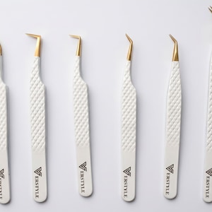 May include: A set of six tweezers with white textured handles and gold-colored tips. Each tool features a unique angled or curved head design and the black text ERSTYLE printed near the base of the handle, arranged in a vertical row.