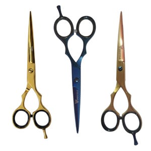 May include: Three professional hair cutting shears arranged vertically. The left and right shears are gold-toned with black finger rings. The center shear is dark blue with a metallic finish. Each blade is engraved with the brand name VERSTYLE.