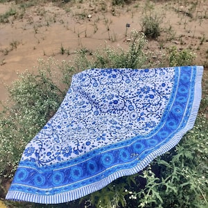 May include: A blue and white floral patterned textile, possibly a tablecloth or throw, is displayed outdoors. The design features a central floral motif with a border of repeating circular patterns and stripes. The textile is draped over greenery.