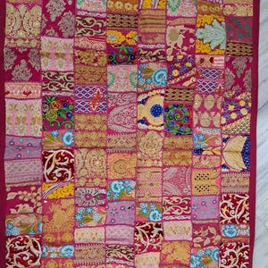 Puede incluir: Textil de retazos con un borde rosa oscuro y una cuadrícula de cuadrados estampados variados. Los cuadrados presentan diseños ornamentados en tonos de rosa, dorado, azul y verde. La estética general es rica y detallada, lo que sugiere un artículo hecho a mano.