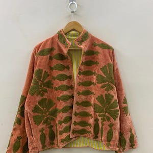 May include: A coral-colored jacket with olive green embroidered floral and leaf patterns. The jacket has a mandarin collar and a yellow and white striped lining. It is hanging on a wooden hanger against a white wall.