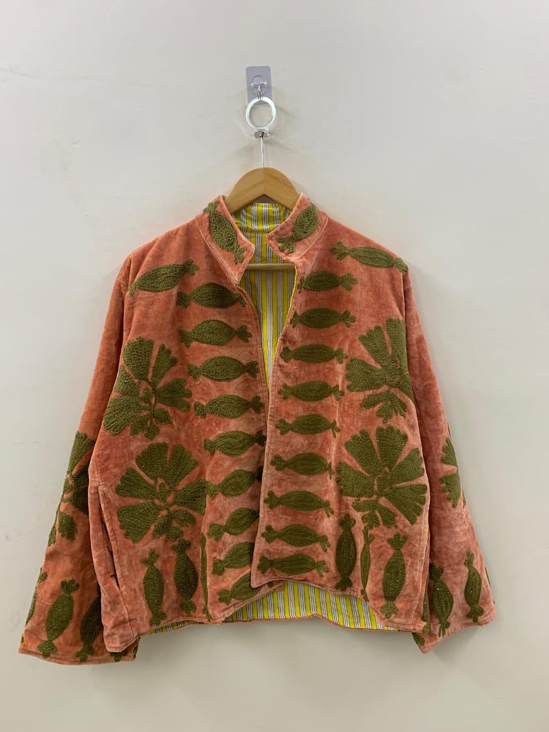 May include: A coral-colored jacket with olive green embroidered floral and leaf patterns. The jacket has a mandarin collar and a yellow and white striped lining. It is hanging on a wooden hanger against a white wall.