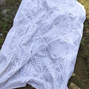 May include: White fabric with a cut-out floral pattern. The fabric is draped over a surface, with the pattern visible across the surface. The fabric has a textured appearance and is likely made of cotton or a similar material. The text "Shot on OnePlus SellerTextileExport" is visible in the bottom left corner.