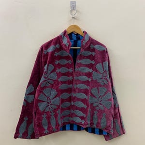 May include: A burgundy velvet jacket with gray embroidered floral and abstract designs. The jacket has a mandarin collar and long sleeves. The interior lining is blue with vertical stripes. The jacket is hanging on a wooden hanger.