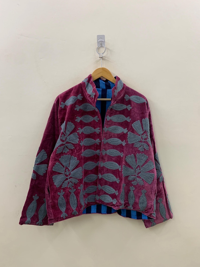 May include: A burgundy velvet jacket with gray embroidered floral and abstract designs. The jacket has a mandarin collar and long sleeves. The interior lining is blue with vertical stripes. The jacket is hanging on a wooden hanger.