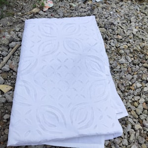 May include: A folded white fabric with a repeating floral pattern. The fabric is made of a soft material and has a delicate, openwork design. The fabric is lying on a bed of gray rocks.