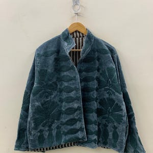 May include: A teal velvet jacket with dark green floral embroidery. The jacket has a high collar and a striped lining. It is hanging on a wooden hanger against a neutral wall.