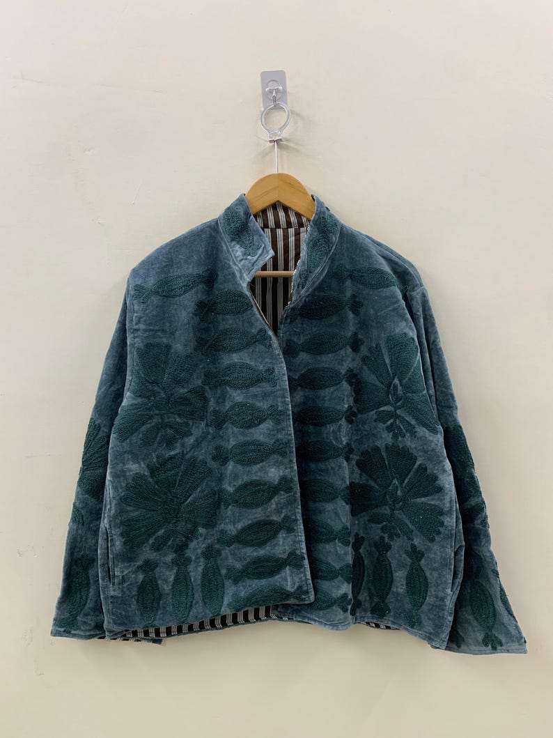 May include: A teal velvet jacket with dark green floral embroidery. The jacket has a high collar and a striped lining. It is hanging on a wooden hanger against a neutral wall.